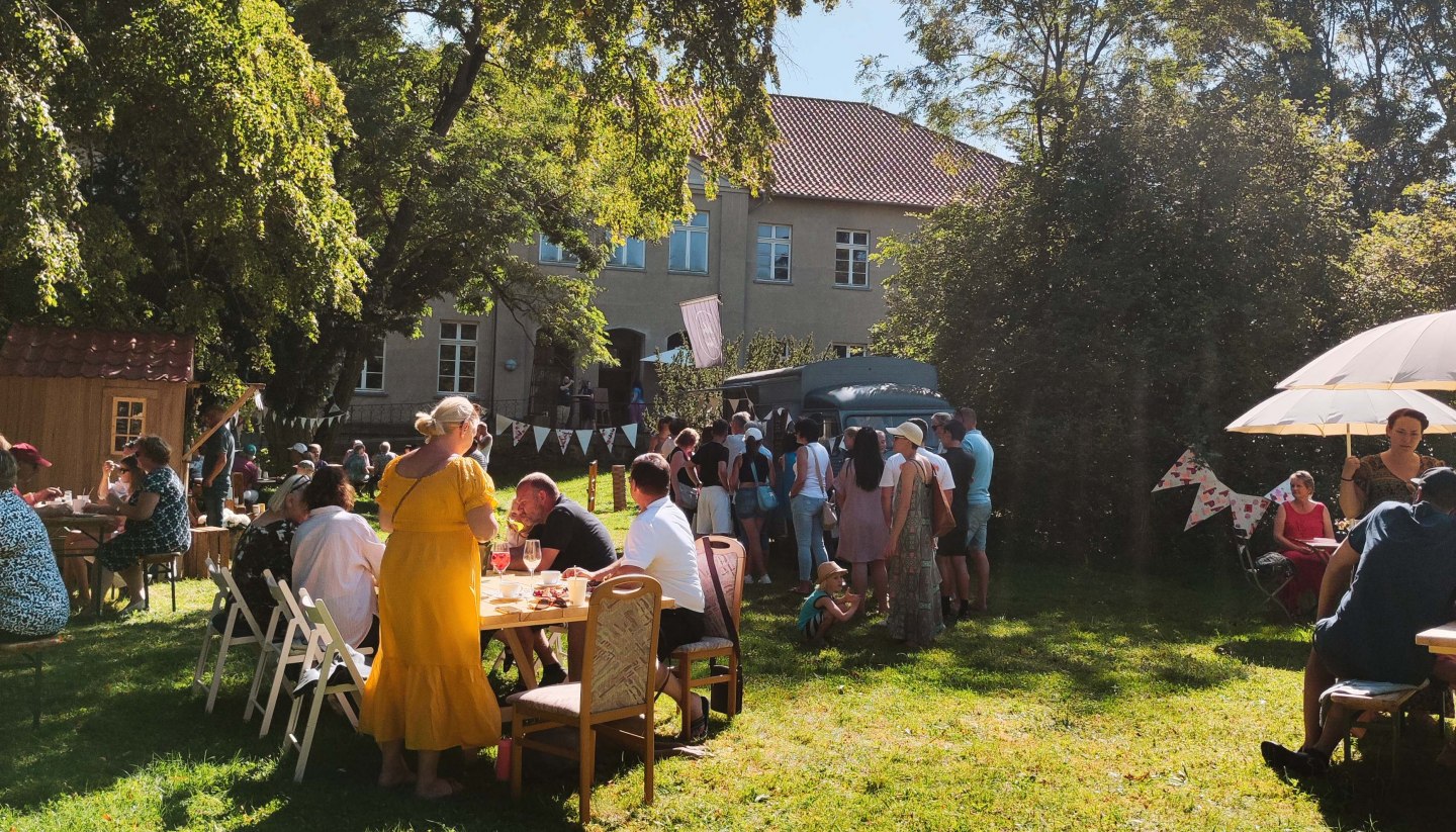 The MitsommerRemise in Klocksin Manor Park with food and drinks. People sit at tables in the park behind the manor house.