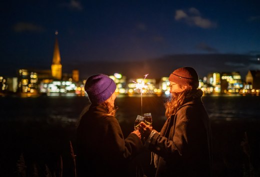 Two people toast with sparkling wine while holding a sparkler, with the illuminated skyline of Rostock in Mecklenburg-Vorpommern in the background.