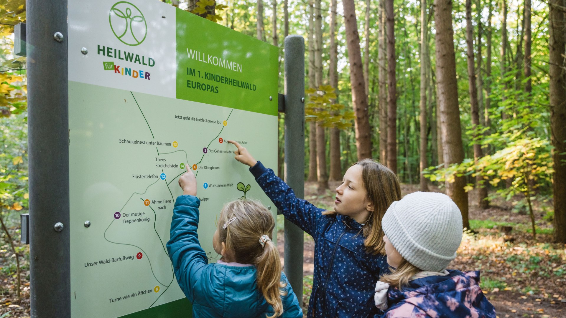 Three children stand in front of a large overview map in the forest, point to various points with their fingers and plan their route.