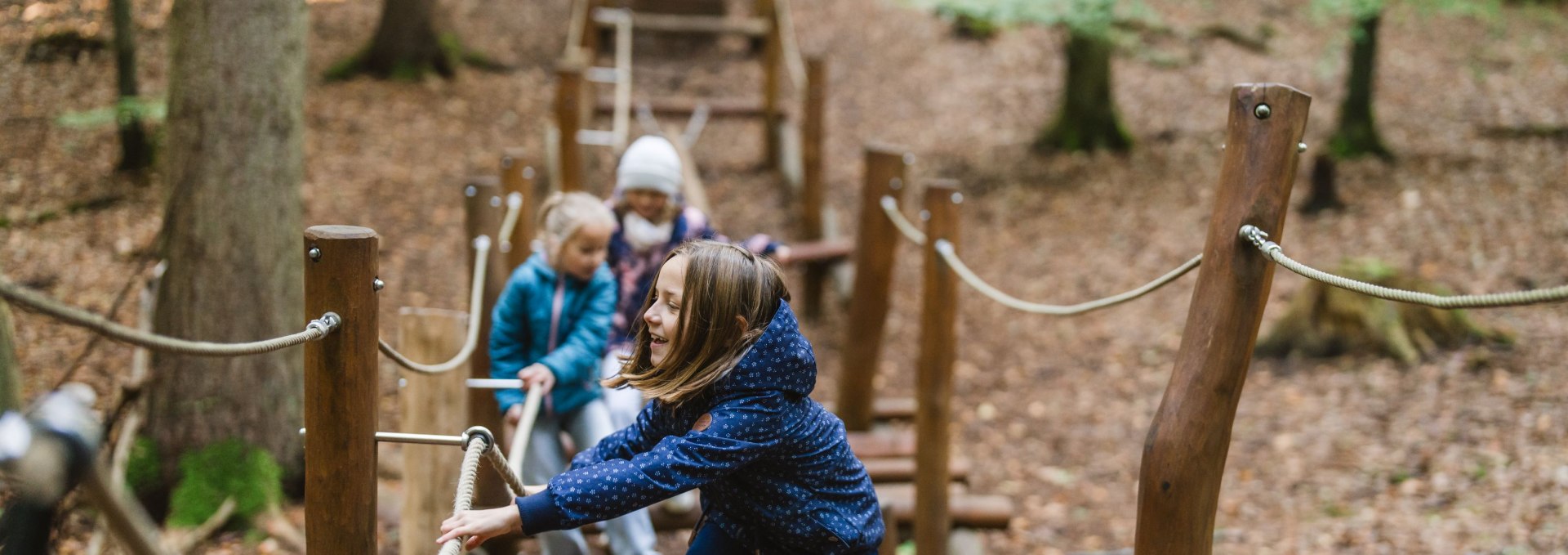 A child balances on a narrow beam on a forest playground while two other children run along the course in the background.