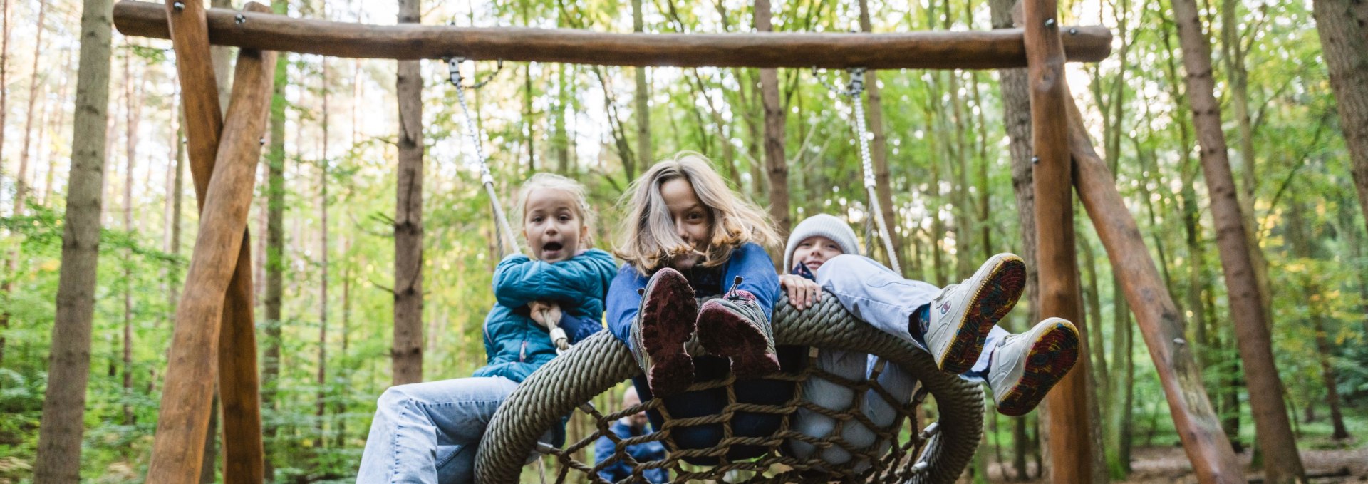 Three children sit together on a nest swing made of ropes in the forest as they swing wildly.