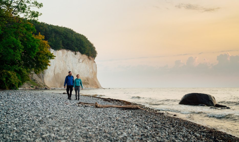 A couple walks along the foot of the famous chalk cliffs on the island of Rügen, while the sunset lights up the sky in soft colors.