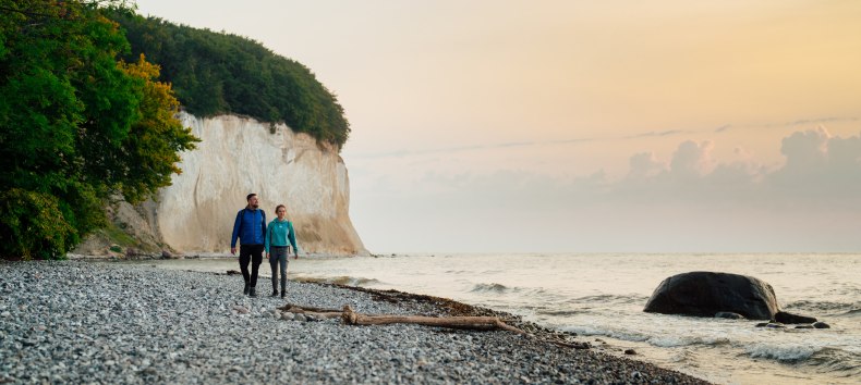 A couple walks along the foot of the famous chalk cliffs on the island of Rügen, while the sunset lights up the sky in soft colors.