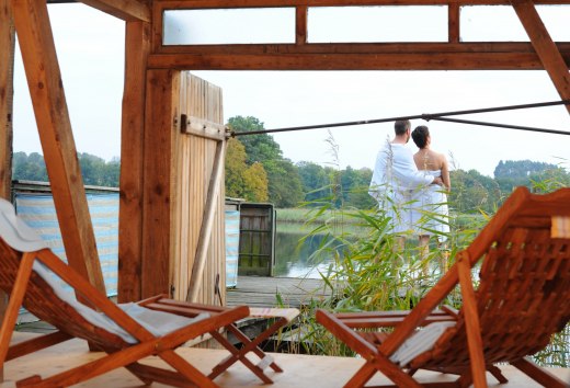 A couple in towels stands on a terrace by the lake, surrounded by reeds, with a view of the water in the Feldberg lake landscape. Wooden loungers can be seen in the foreground.