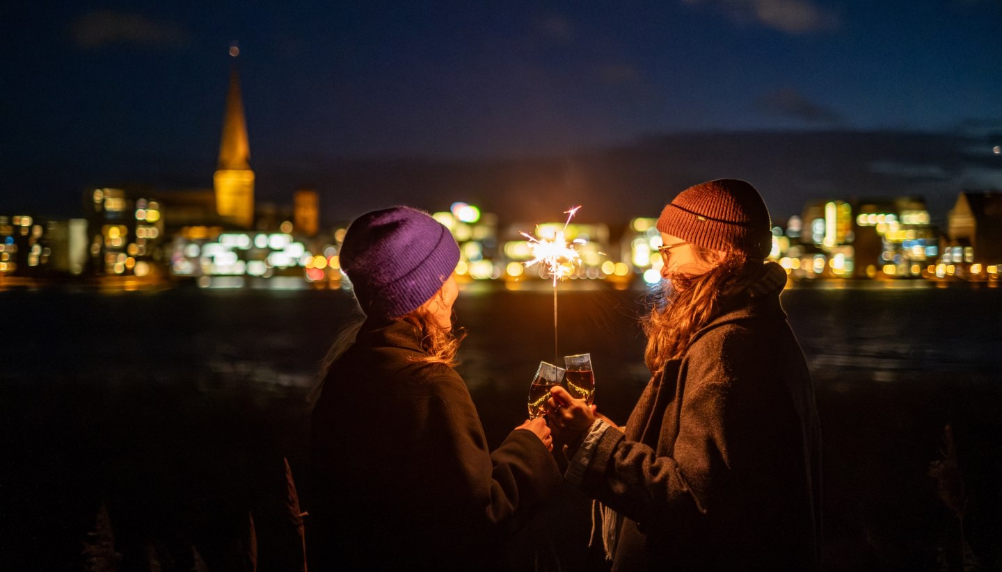 Two people toast with sparkling wine while holding a sparkler, with the illuminated skyline of Rostock in Mecklenburg-Vorpommern in the background.