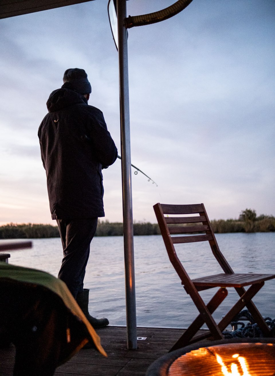 Person fishing from the deck of a houseboat on the Peene at sunset, with a fireplace in the foreground.