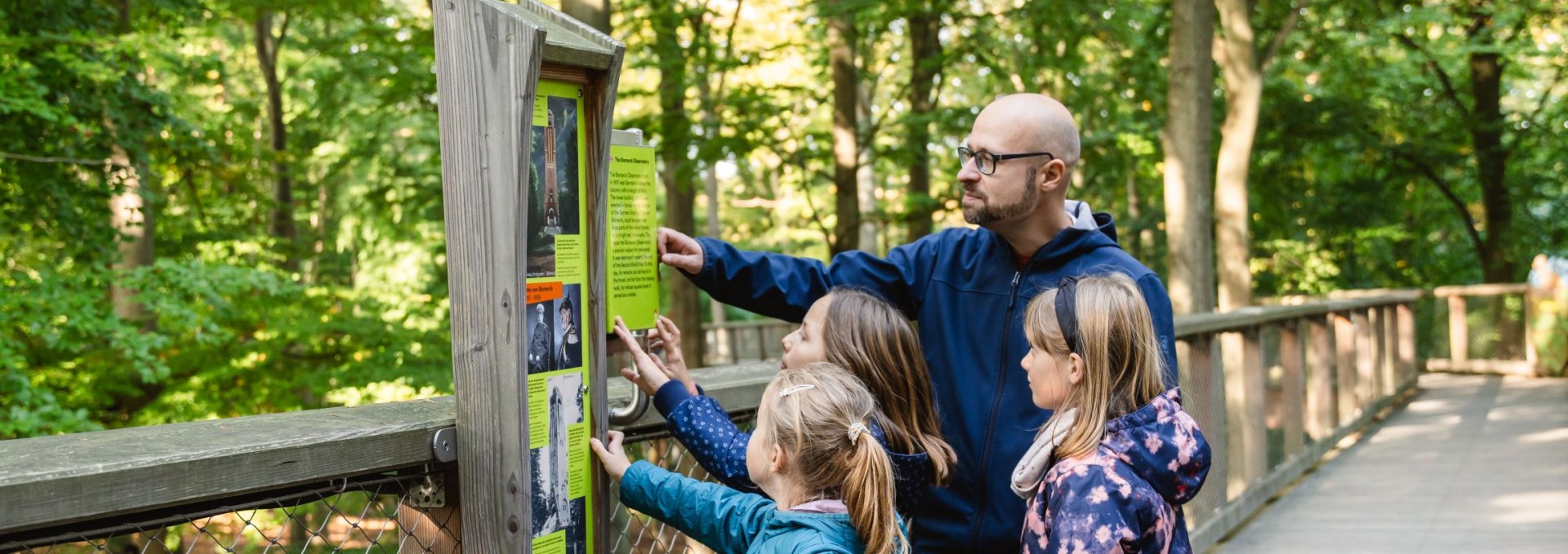 A group of children and an adult look at an information board on a wooden treetop path surrounded by green trees.