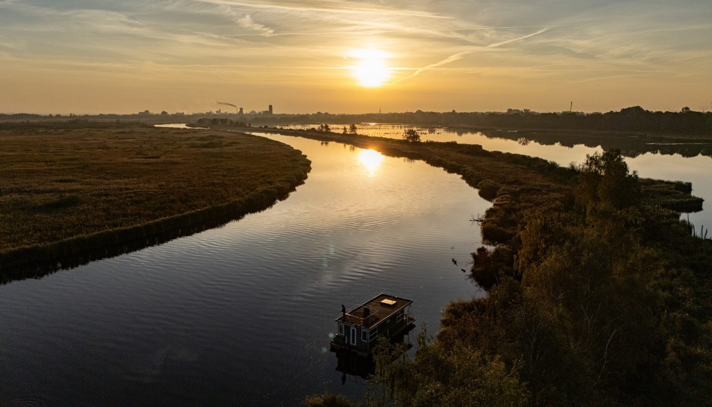 Houseboat on the Peene at sunset, surrounded by a tranquil river landscape and golden light on the horizon.