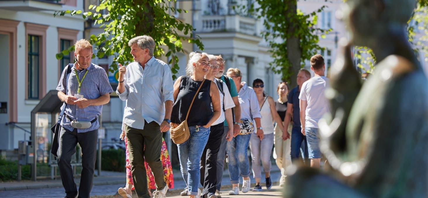 A group of people are walking along a cobbled path with a city guide. On the right, blurred in the picture, a statue can be seen by the Pfaffenteich pond; the villas appear in the background. The focus is on the guide and a couple having a lively conversation., © Oliver Borchert