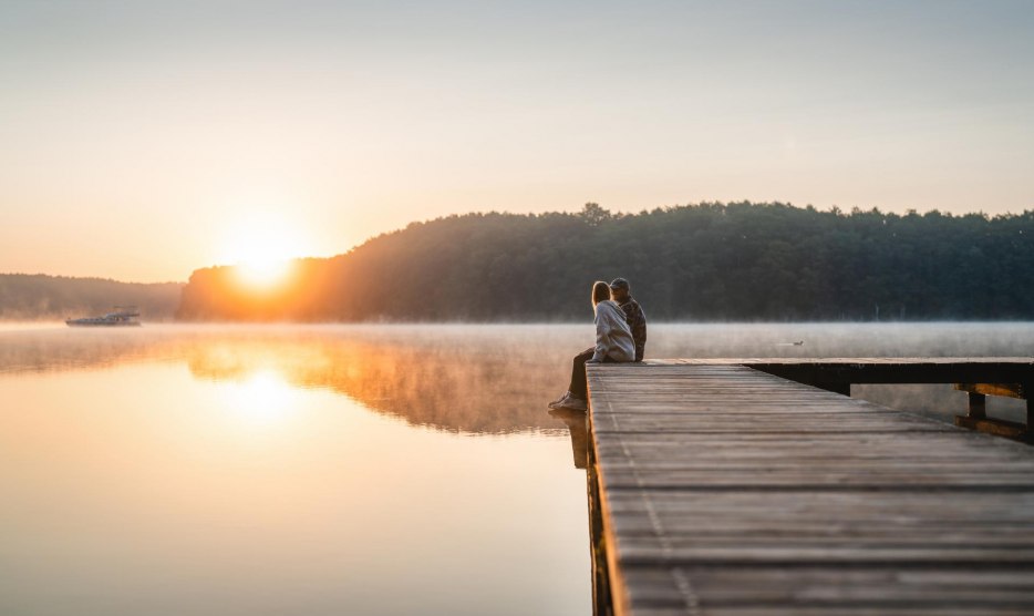 A couple sits on a jetty at sunset on Lake Mirov. A houseboat passes by in the background.