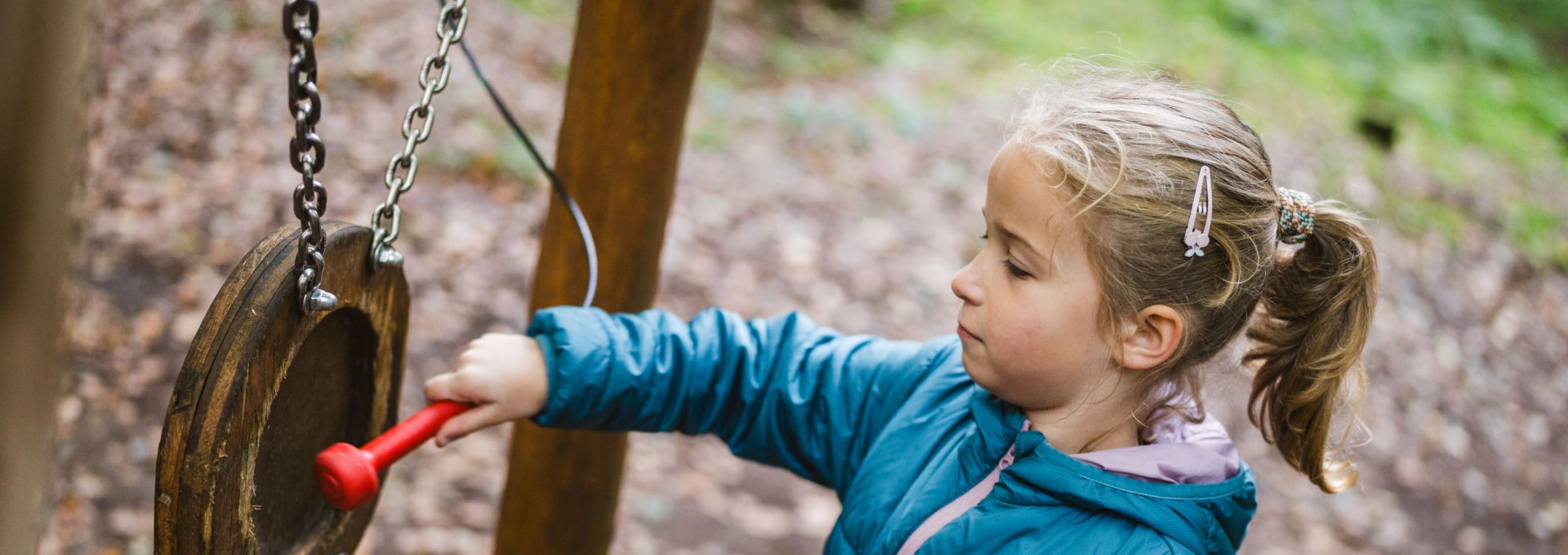 A young girl in a blue jacket strikes a wooden sound station with a red mallet, surrounded by trees and forest.