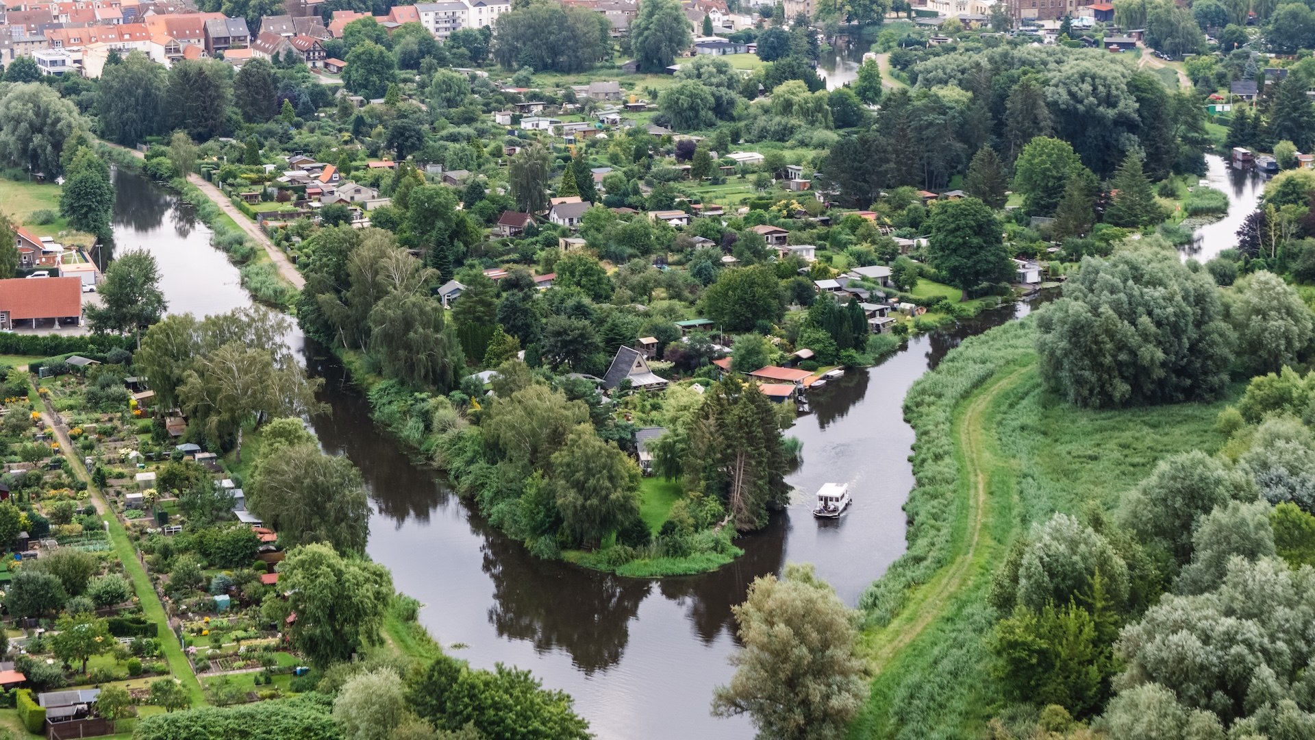 An aerial view shows the winding Elde with a houseboat surrounded by green gardens and the town of Parchim in the background.