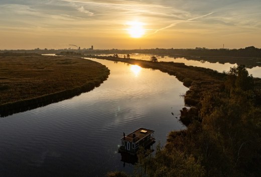 Houseboat on the Peene at sunset, surrounded by a tranquil river landscape and golden light on the horizon.