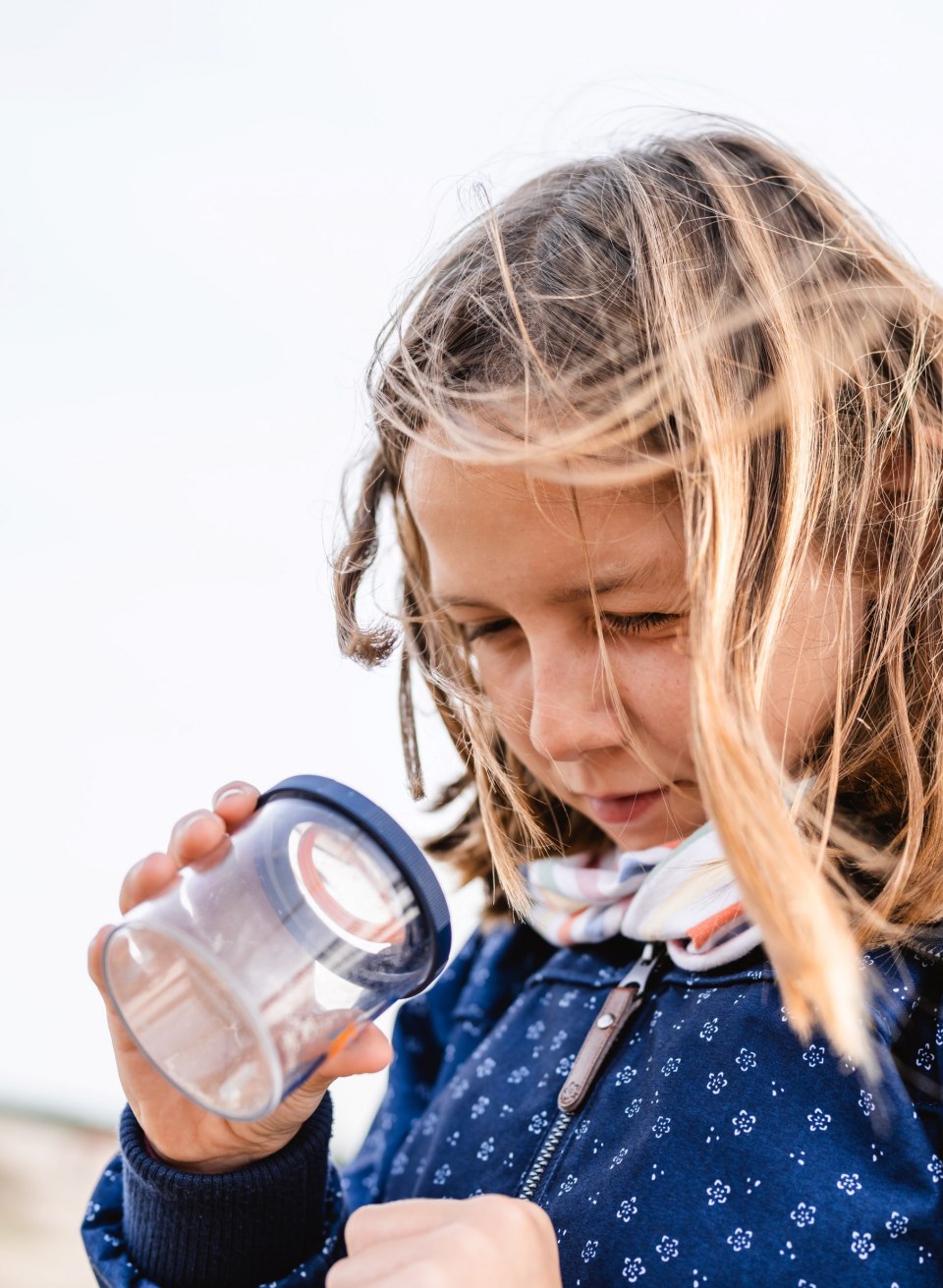 A girl with tousled hair holds a magnifying glass and looks intently at the contents while the wind blows through her hair.