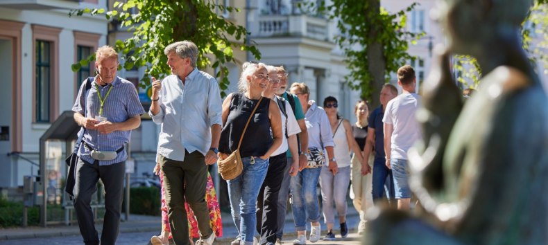 A group of people are walking along a cobbled path with a city guide. On the right, blurred in the picture, a statue can be seen by the Pfaffenteich pond; the villas appear in the background. The focus is on the guide and a couple having a lively conversation., © Oliver Borchert