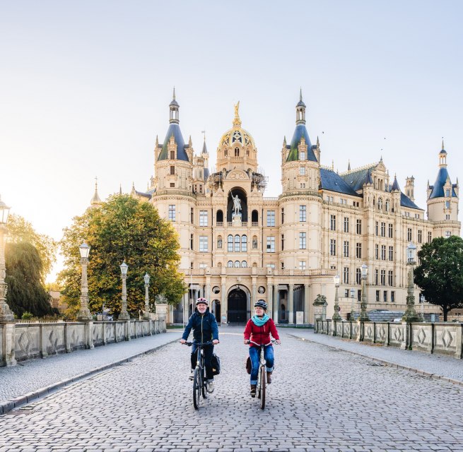 Two cyclists cross a bridge in front of Schwerin Castle, illuminated by the golden morning sun.