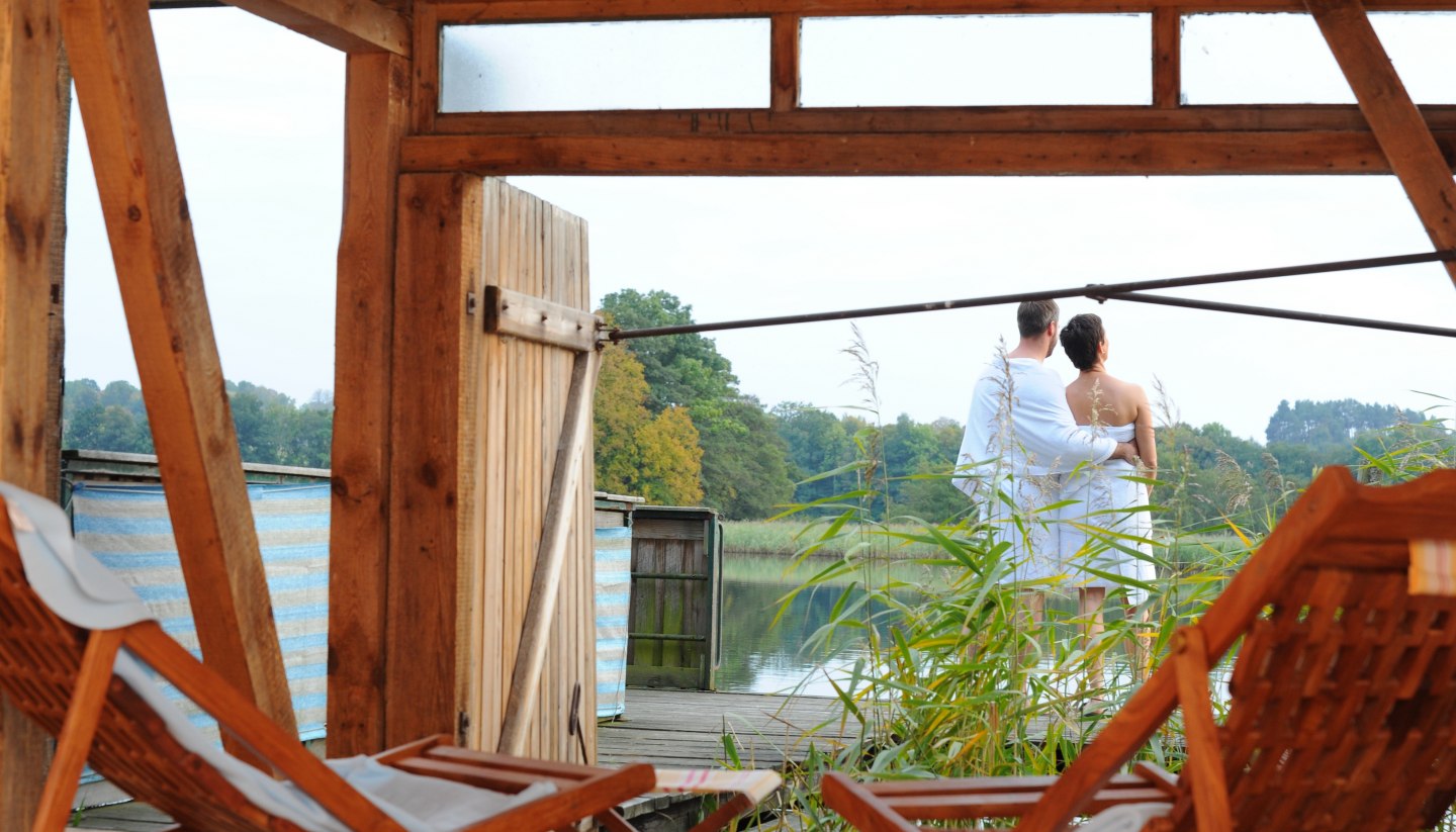 A couple in towels stands on a terrace by the lake, surrounded by reeds, with a view of the water in the Feldberg lake landscape. Wooden loungers can be seen in the foreground.