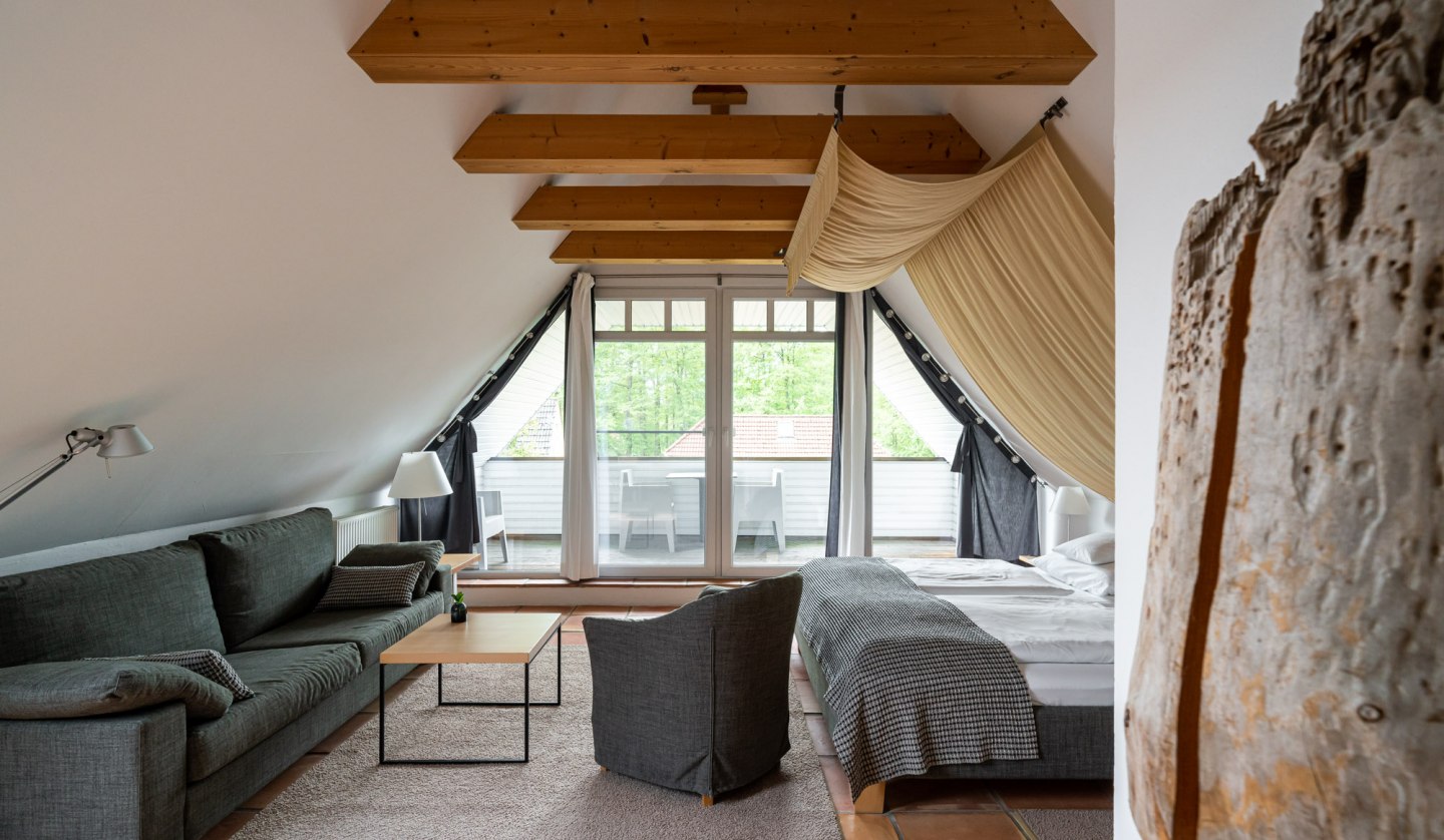 A stylishly furnished attic room with exposed wooden beams and a cozy ambience. The room offers a comfortable seating area, a large bed and access to a balcony with a view of the greenery. Warm terracotta floors and light fabrics round off the cozy design and invite you to relax., © Hotel Carpe Diem