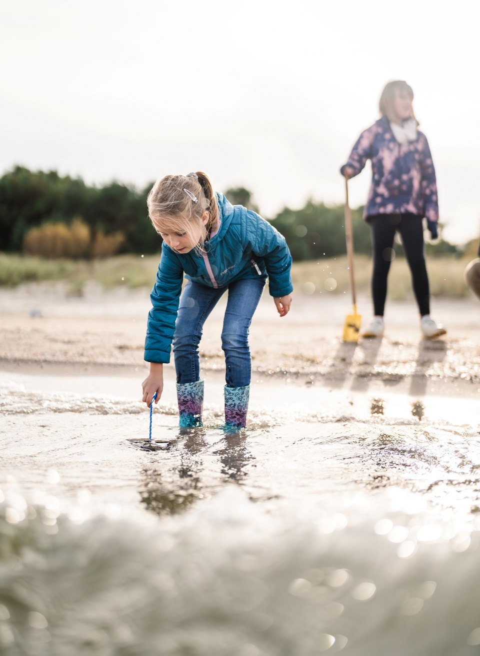 A girl in rubber boots stands bent over in the water on the beach, looking for something in the sand, while another child stands in the background.