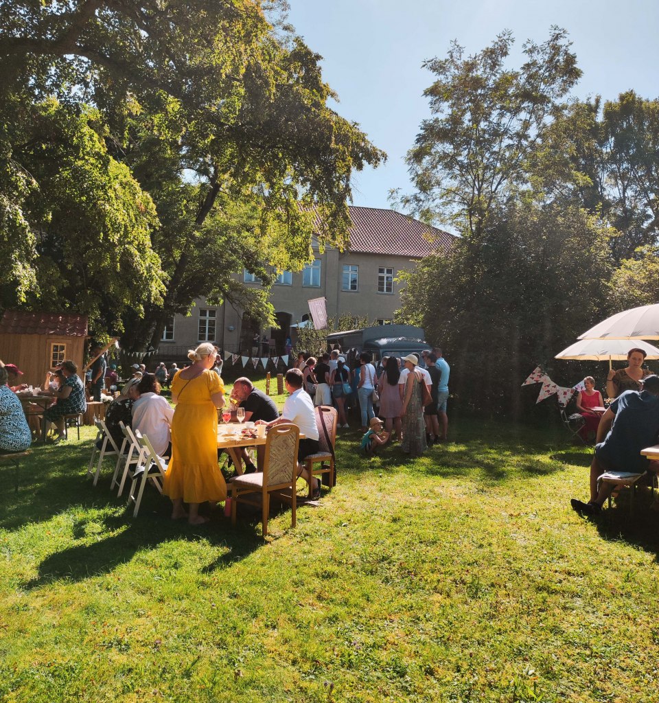 The MitsommerRemise in Klocksin Manor Park with food and drinks. People sit at tables in the park behind the manor house.