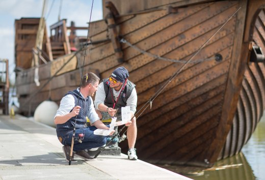 Young men fishing at the town harbor in Ueckermünde with a cog in the background., © TMV/Läufer