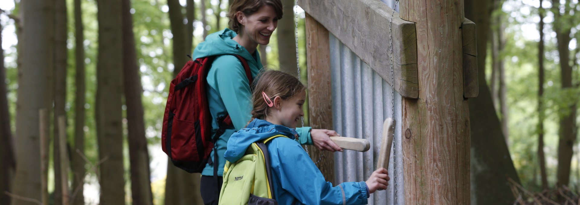 A mother and her daughter stand in front of an installation with sound tubes in the sound forest in Graal-Müritz. Both are holding wooden mallets and striking the tubes to produce sounds while smiling.