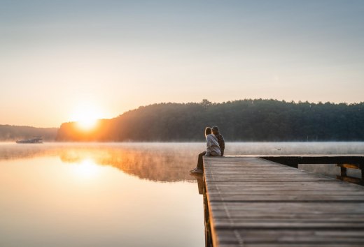 A couple sits on a jetty at sunset on Lake Mirov. A houseboat passes by in the background.