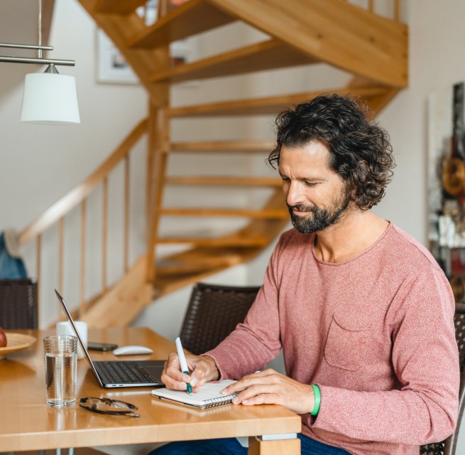 A man sits at a wooden table in a vacation home, working on his laptop and taking notes - ideal conditions for a workation on the island of Poel.