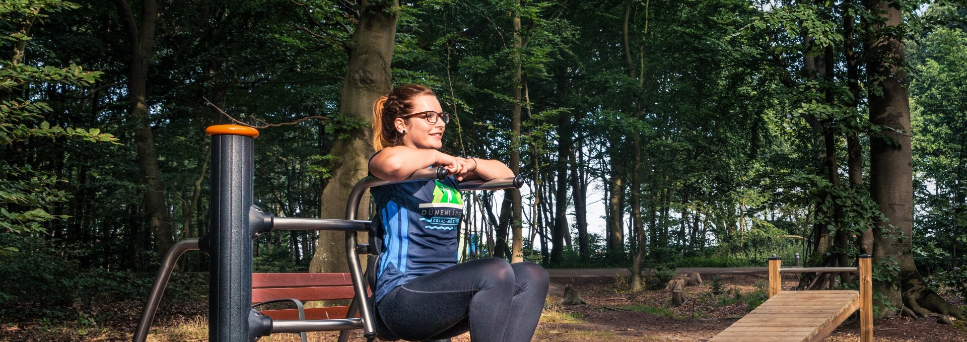 A woman works out on a fitness machine in the Graal-Müritz active forest, surrounded by tall trees and a natural forest atmosphere.