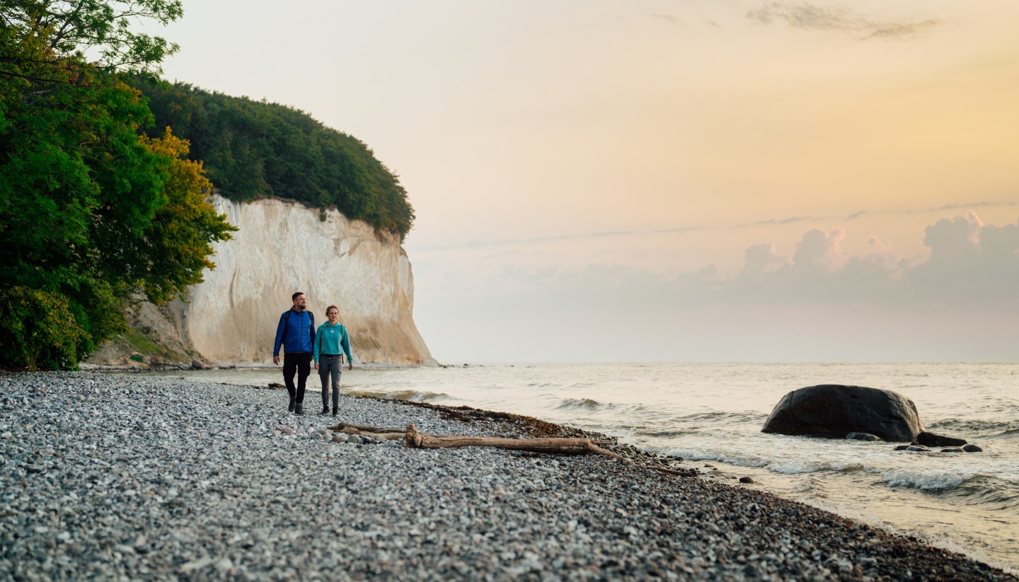 A couple walks along the foot of the famous chalk cliffs on the island of Rügen, while the sunset lights up the sky in soft colors.