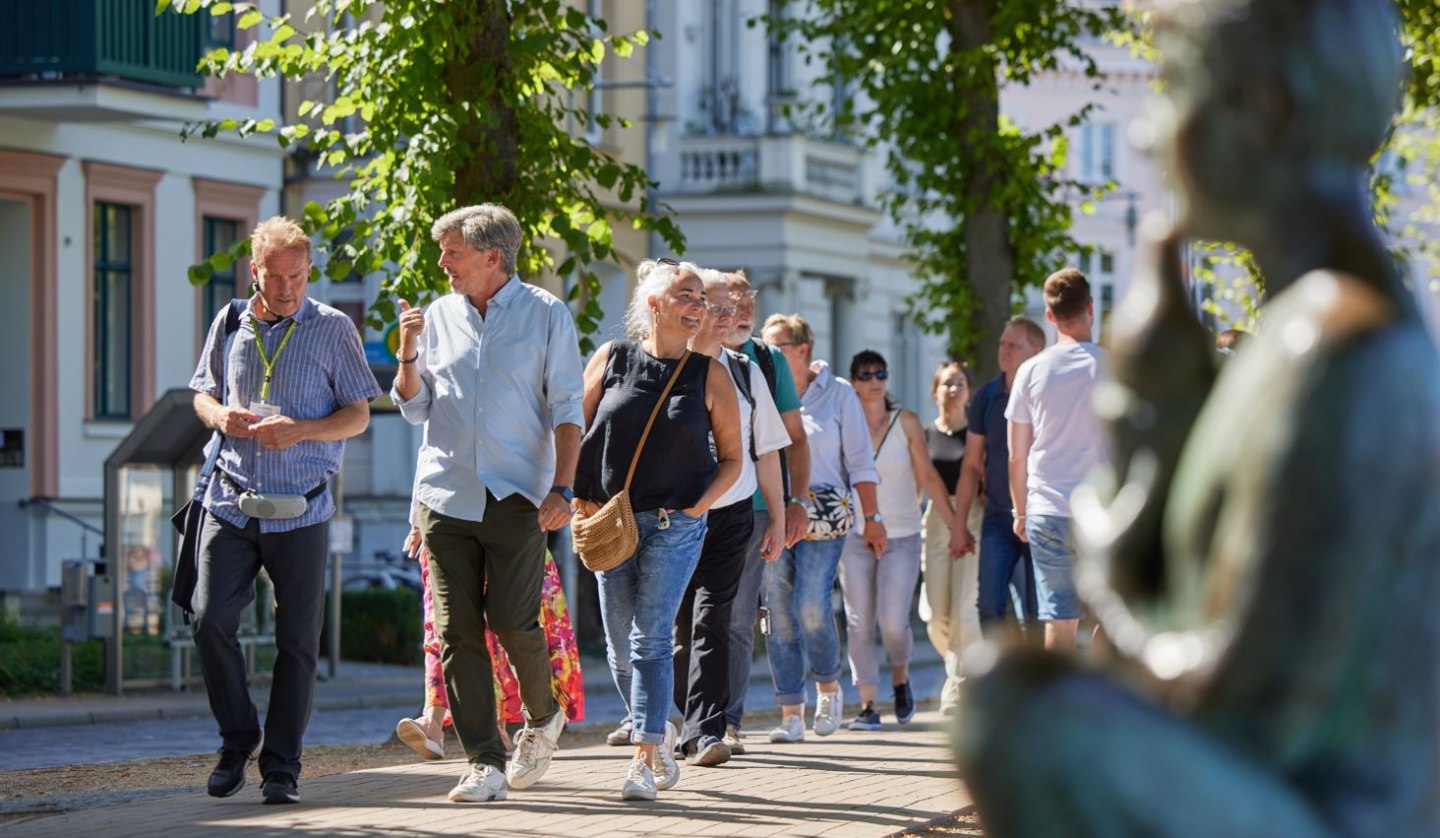 A group of people are walking along a cobbled path with a city guide. On the right, blurred in the picture, a statue can be seen by the Pfaffenteich pond; the villas appear in the background. The focus is on the guide and a couple having a lively conversation., © Oliver Borchert