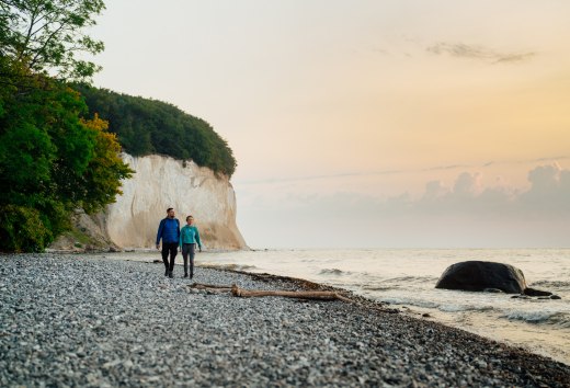 A couple walks along the foot of the famous chalk cliffs on the island of Rügen, while the sunset lights up the sky in soft colors.