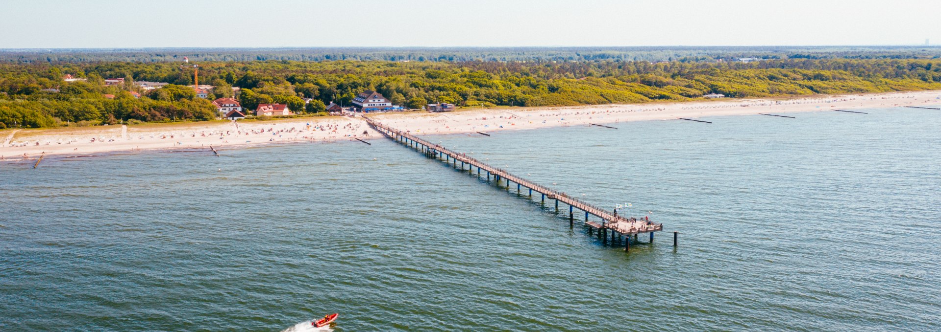 Aerial view of the pier in the Baltic Sea spa town of Graal-Müritz, which juts far out into the Baltic Sea, surrounded by a sandy beach and dense green forests in the background.