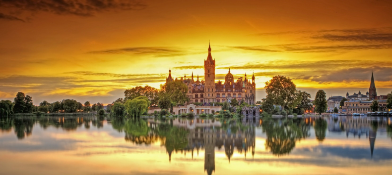 Schwerin Castle is framed by the last rays of the sun. The sunset colors the sky orange. Schwerin Castle is reflected in the calm lake.