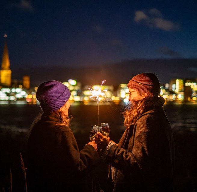 Two people toast with sparkling wine while holding a sparkler, with the illuminated skyline of Rostock in Mecklenburg-Vorpommern in the background.