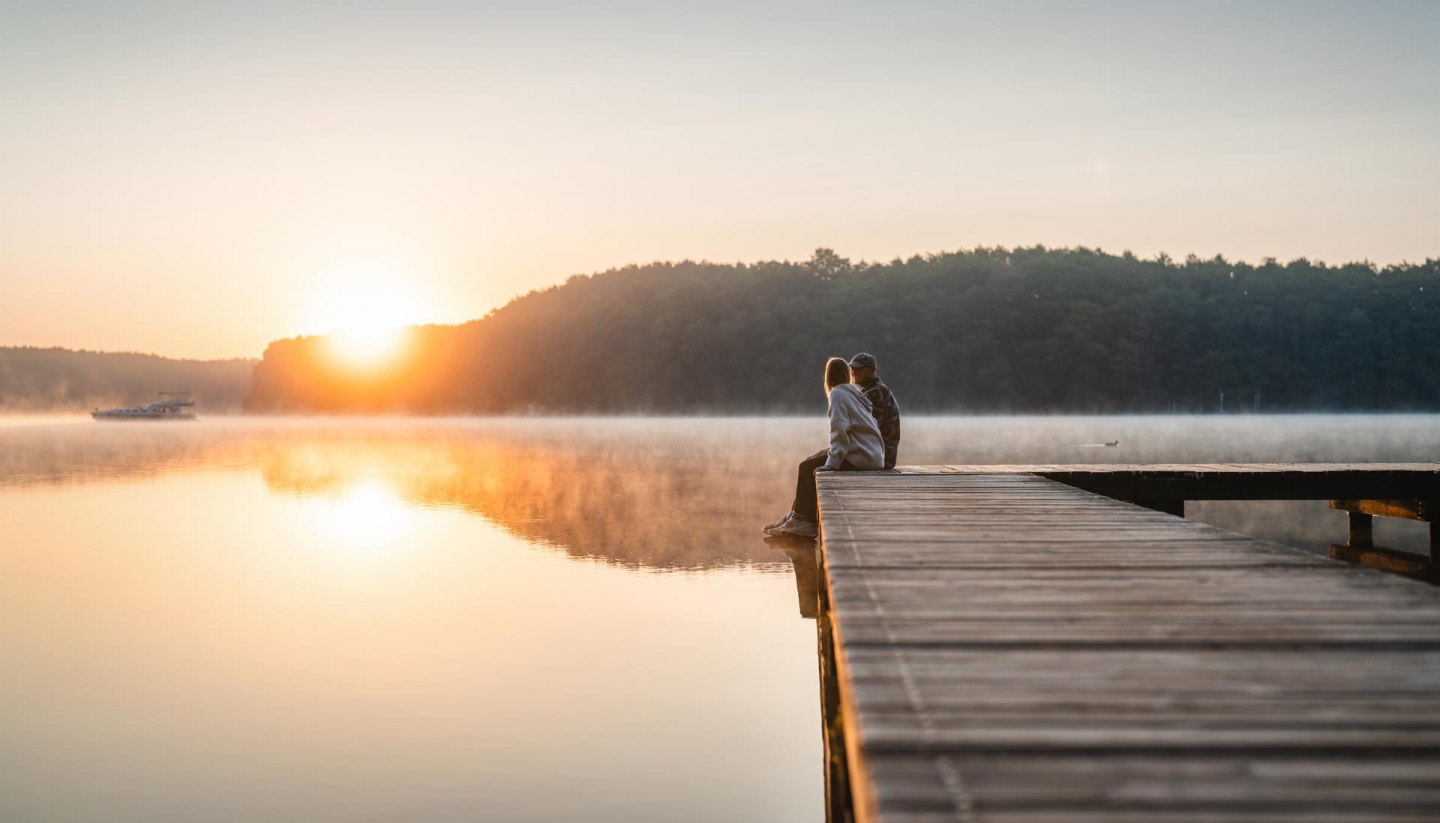 A couple sits on a jetty at sunset on Lake Mirov. A houseboat passes by in the background.