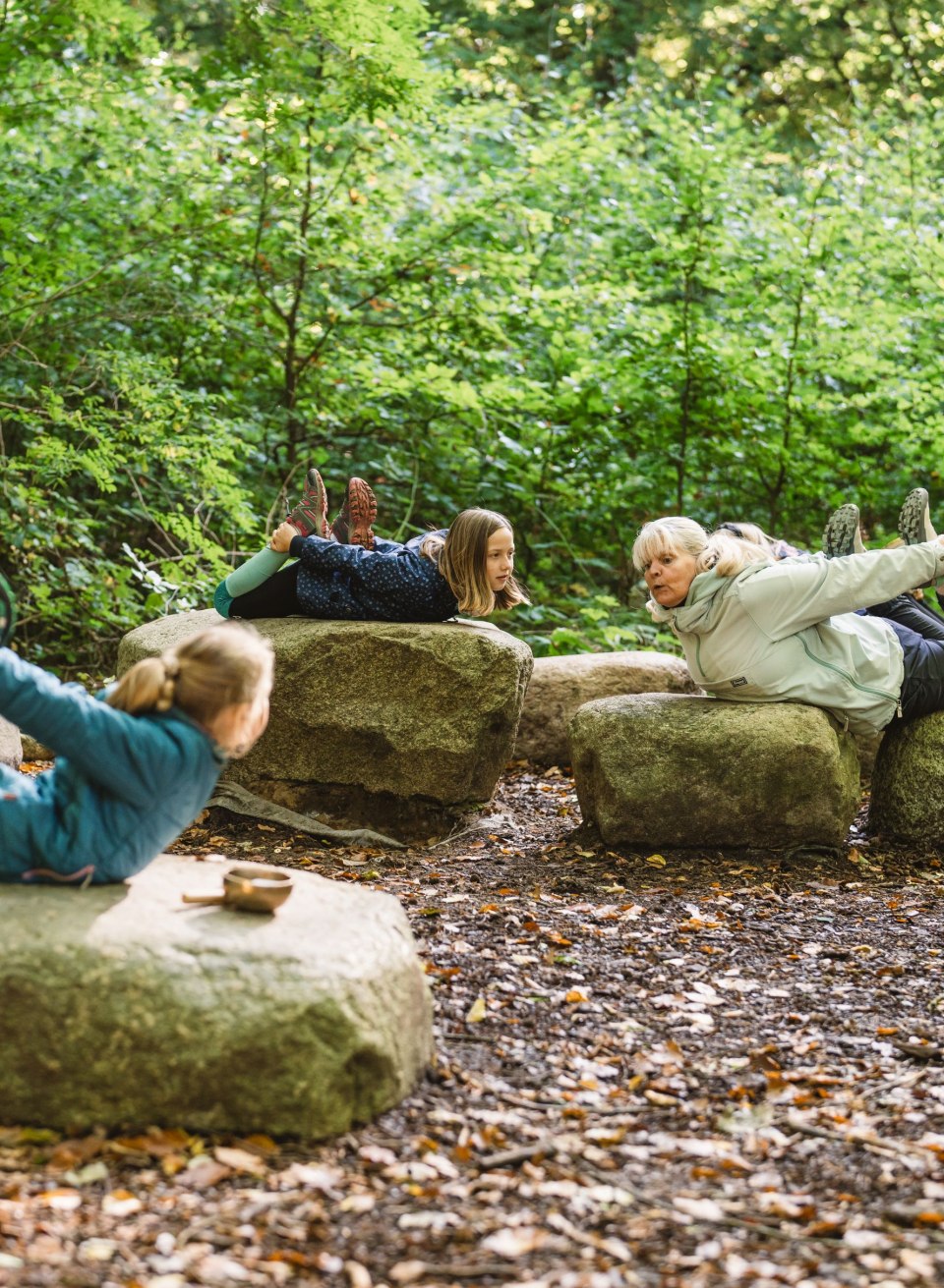 Children and an adult lie on large stones in the forest and do yoga exercises together, surrounded by dense trees and leaves.