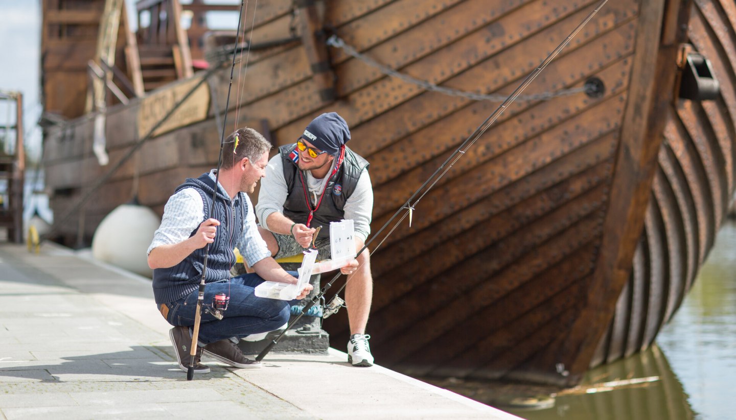 Young men fishing at the town harbor in Ueckermünde with a cog in the background., © TMV/Läufer