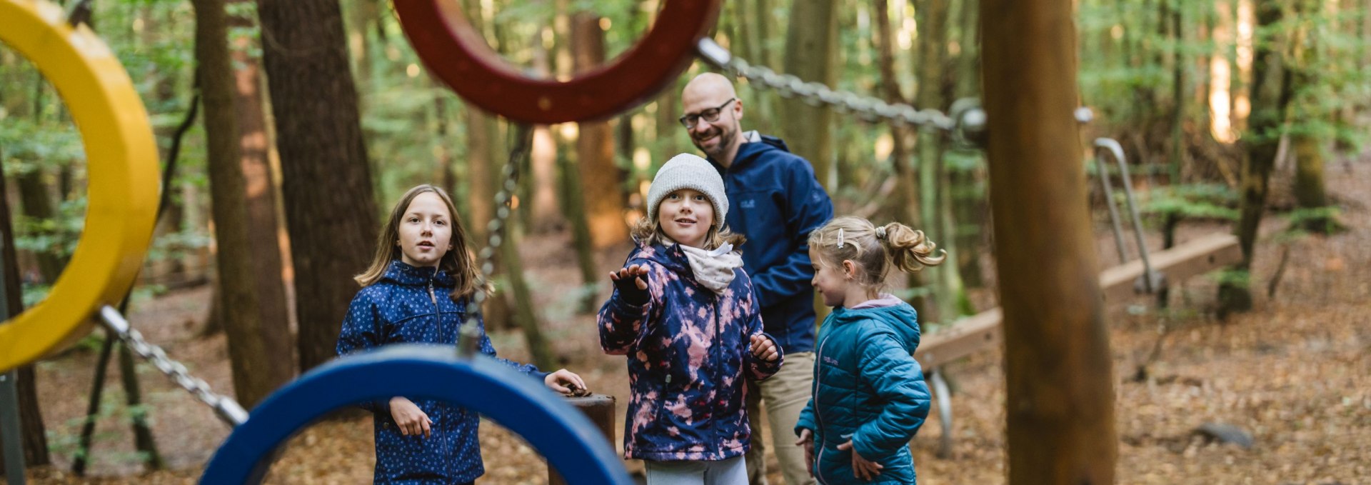 A group of children and an adult stand between colorful metal climbing rings on a forest playground.