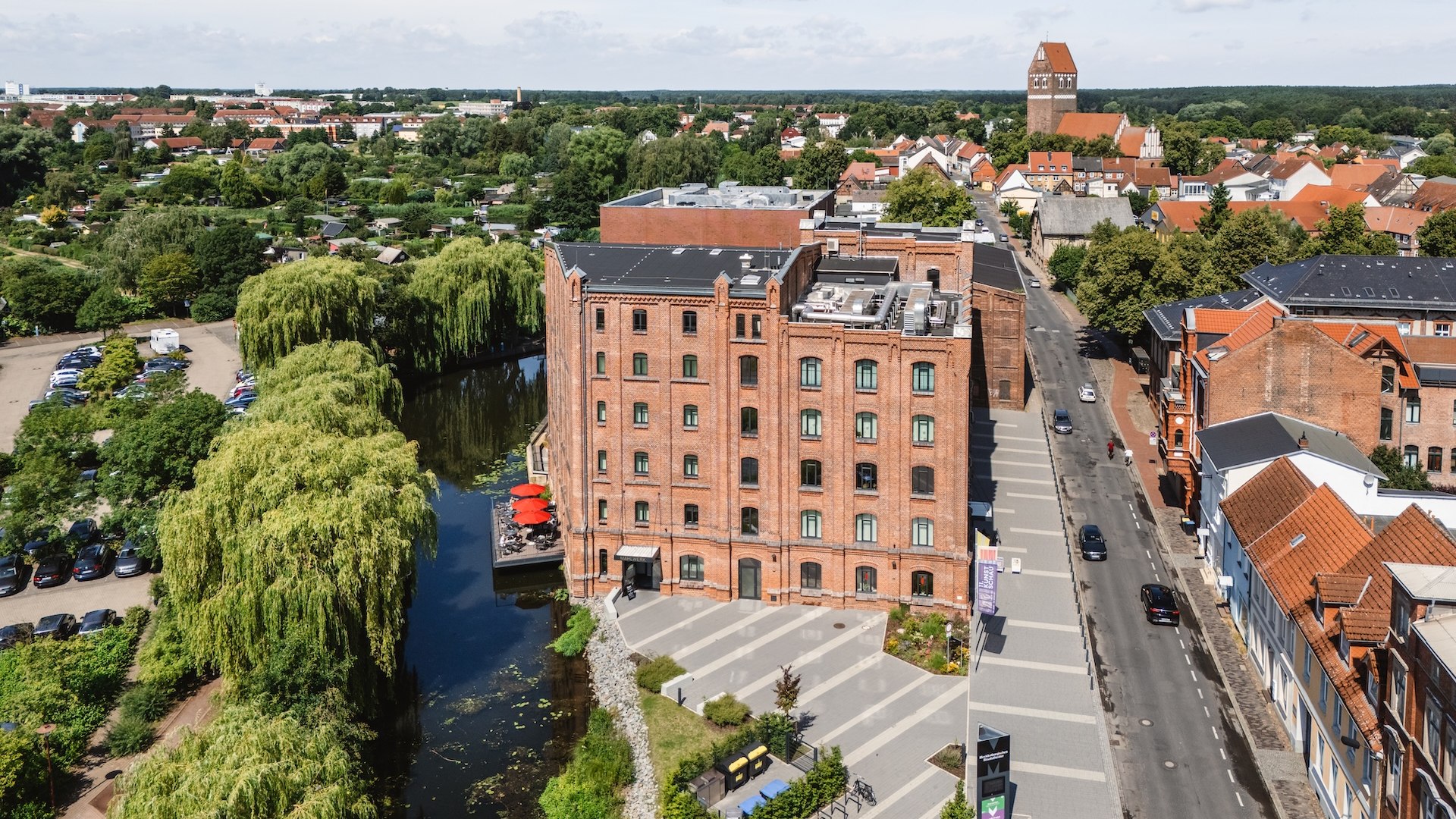 An aerial view shows the Kulturmühle in Parchim with its historic façade and a cozy restaurant terrace by the water, surrounded by greenery.