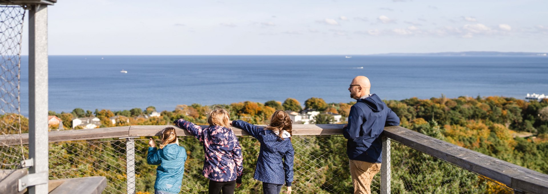  Two adults and three children stand on a viewing platform and look out over the forest to the blue sea.