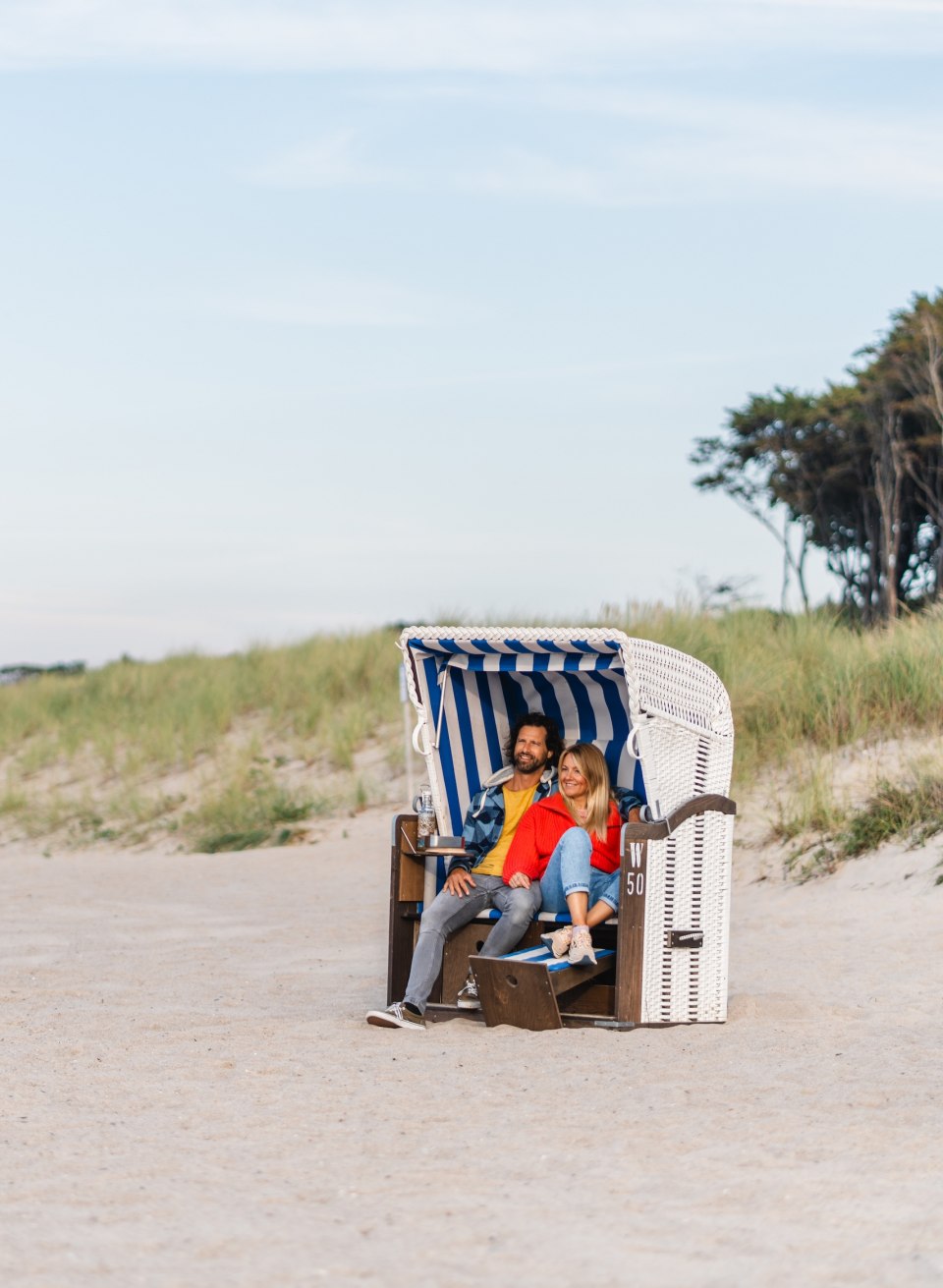 A couple sits in a blue and white striped beach chair on the beach in Graal-Müritz, surrounded by sand and dunes with a view of the tranquil coastal landscape.