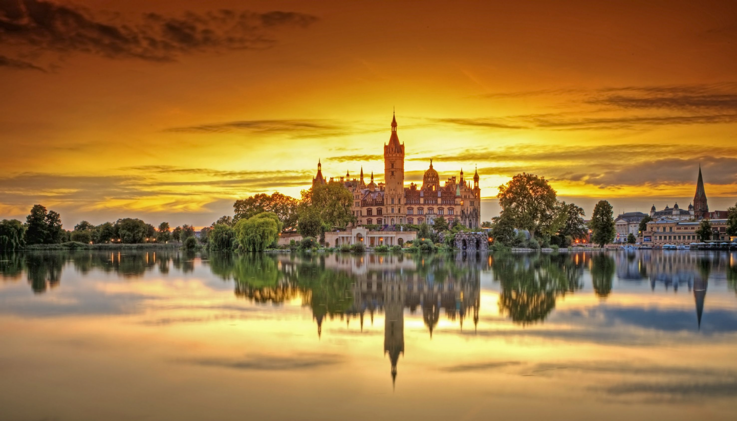 Schwerin Castle is framed by the last rays of the sun. The sunset colors the sky orange. Schwerin Castle is reflected in the calm lake.