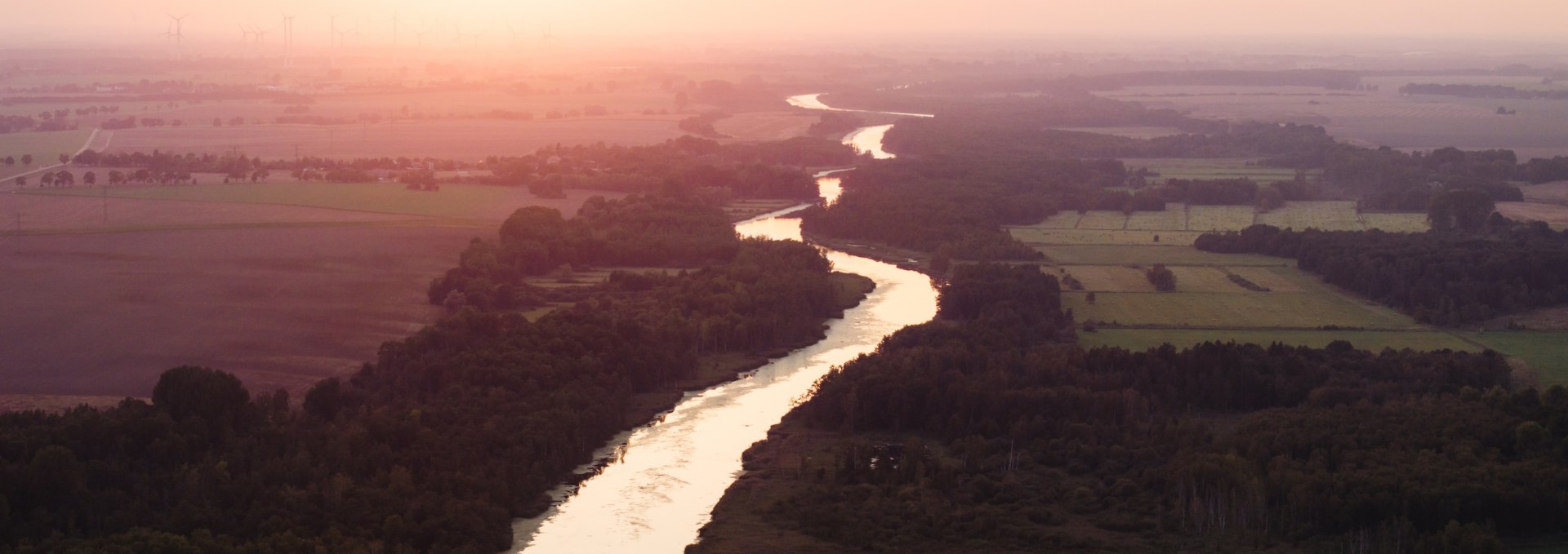 Aerial view of the Peene at sunset, embedded in fields and forests, with an orange sky in the background.